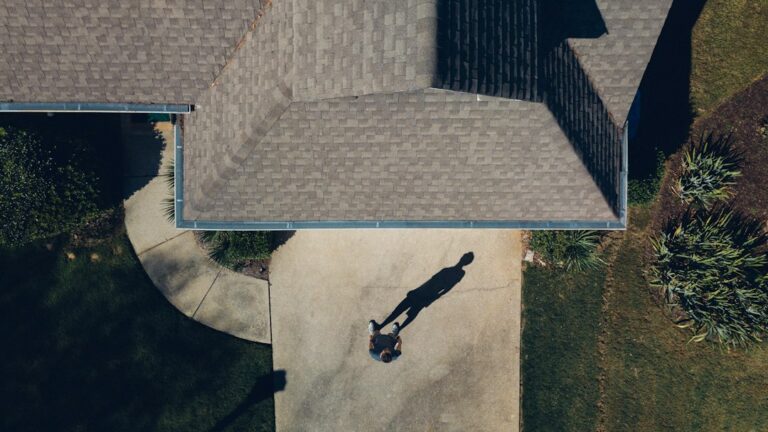 A homeowner examines roof shingles and gutters from the ground while holding a flashlight and inspection notes.
